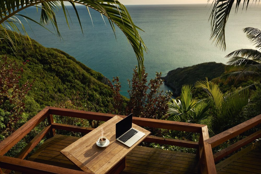 Arbeitsplatz mit Laptop auf einer Holzterrasse über einer tropischen Bucht (KI-generiertes Symbolbild)